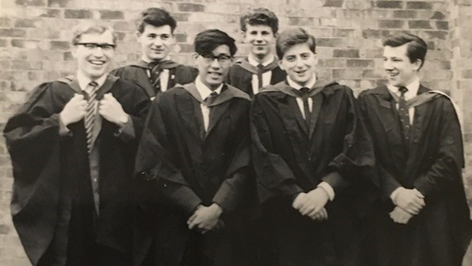 John McGrail stands with a group of five friends. They wear gowns and are standing in front of a brick wall - which is Faraday Hall. 